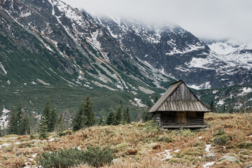 Fototapeta na wymiar Old wooden cottage among high mountains, fiords landscape, with fog and snow all around. Concept of isolation, social distance travel, trekking in Europe. Moody, desaturated colors.