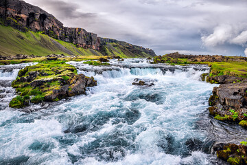 Landscape view of rushing river by Fjadrargljufur canyon in Iceland with rapids stream of water by green grass moss and cloudy sky