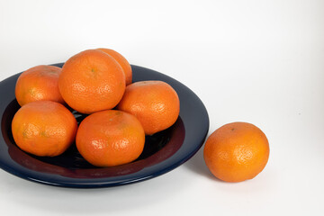 Six mandarin oranges are piled on a dark blue glass plate with one extra orange next to it against a white background.