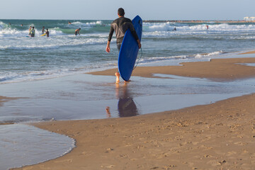 Young surfer with leg rope walking on the beach. Sunny day and big wave, paradise of surfer.