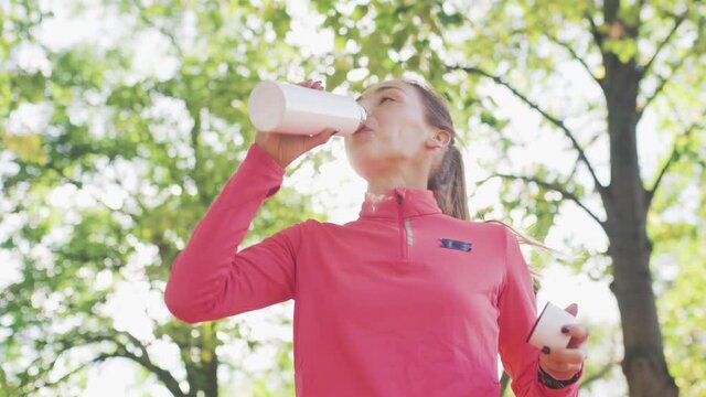 Fitness girl in pink sportswear running and drinking water from a bidon in the park