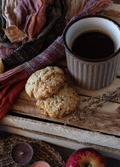 Oatmeal cookies with a mug of black coffee on a wooden box with dried flowers, an apple, candles, an orange-brown scarf