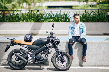 Fototapeta na wymiar Handsome young Asian man sitting on granite parapet next to his motorcycle and resting after ride