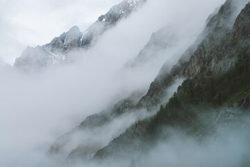 Fototapeta premium Ghostly atmospheric view to big cliff in cloudy sky. Low clouds among giant rocky mountains. Mysterious place at early foggy morning. Minimalist scenery with beautiful rockies. Dramatic bleak fog.