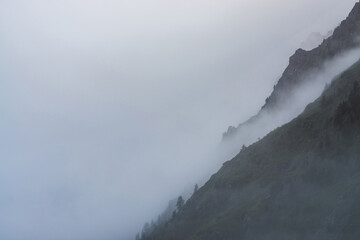 Ghostly atmospheric view to big cliff in cloudy sky. Low clouds among giant rocky mountains. Mysterious place at early foggy morning. Minimalist scenery with beautiful rockies. Dramatic bleak fog.