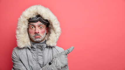 Winter vacation and pastime concept. Surprised red faced man spends much time on frosty weather outside wears grey jacket and hood on head points away at blank space against rosy background.