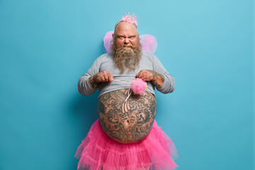 Photo of funny bearded man frowns face and looks at camera wears crown fairy wings dressed in...