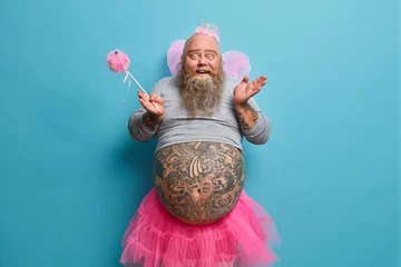 Fotobehang Funny happy bearded man has image of fairy holds magic wand poses with big tattooed belly over blue wall entertains children on party poses against blue background. Adult male dressed like princess © Wayhome Studio