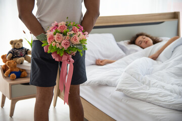 Family relationship and happy anniversary concept. Young romantic man holding the bouquet of flower hiding his back for surprising his wife laying on bed
