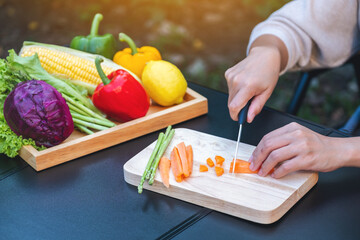 Closeup image of a woman cutting and chopping carrot by knife on wooden board with mixed vegetables in a tray