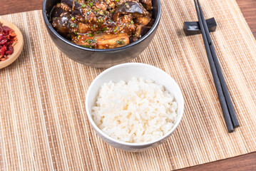 Korean spicy eggplant with spicy sauce and sesame seeds in a bowl and a bowl of boiled rice on a bamboo napkin on a rustic wooden table, top view, close-up