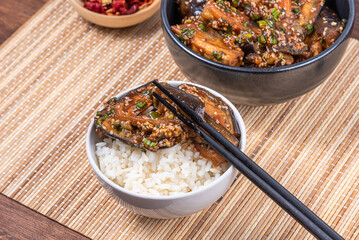 Korean spicy eggplant with spicy sauce and sesame seeds in a bowl and a bowl of boiled rice on a bamboo napkin on a rustic wooden table, top view, close-up