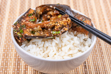 Korean style spicy eggplant with spicy sauce and sesame seeds in a bowl of rice and chopsticks on a bamboo napkin on a rustic wooden table, top view, close-up