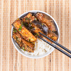 Korean style spicy eggplant with spicy sauce and sesame seeds in a bowl of rice and chopsticks on a bamboo napkin on a rustic wooden table, top view, close-up