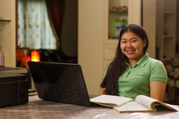 Asian student girl smiling to camera while studying at home, using laptop computer and have a short note on table
