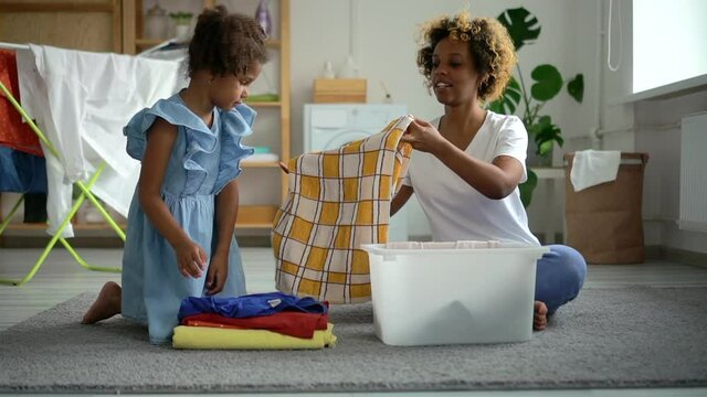 Afro Mother And Daughter Folding Clean Clothes Laundry, Spbd. Mom Teaches Young Kid To Do Housework. Family Help Each Other In Domestic Chores. Stack Of Clothing