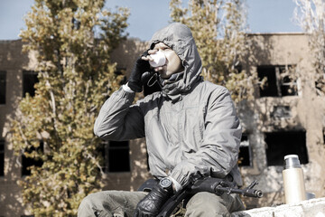 a confident male stalker in a jacket with a hood sits and drinks tea from a thermos, weapons on his lap, against the background of a ruined house