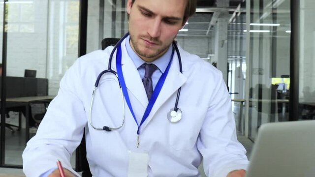 Doctor in white medical uniform with statoscope reading personal file of patient on tablet and writes data to laptop
