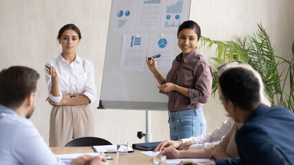 Smiling multiracial female speakers talk interact with employees make whiteboard presentation at...