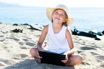 child girl in hat writes draws dreams with sand on seashore