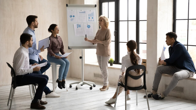 Confident Caucasian middle-aged businesswoman make whiteboard presentation at casual meeting with colleagues. Successful female boss or speaker present business project on flip chart at training.