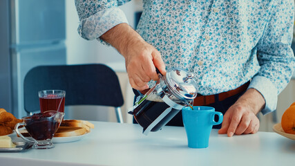 Senior man using french press for coffee preparation and pouring it in mug. Elderly person in the...