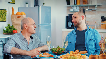 Paralyzed father talking with his son sitting in wheelchair during the family dinner. Happy couples smiling and eating during a gourmet meal, enjoying time sitting around the table in the kitchen