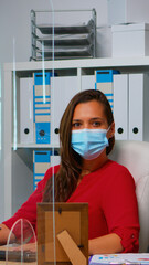 Fototapeta na wymiar Woman with protection mask working and looking at camera sitting in front of pc. Employees in workspace in corporate company typing on computer keyboard looking at desktop respecting social distancing