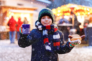 Little cute kid boy eating German sausage and drinking hot children punch on Christmas market....