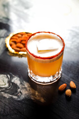 An orange cocktail in a rocks glass with a big ice cube, paprica crust rim, served with almonds