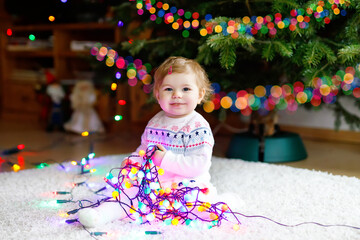 Adorable baby girl holding colorful lights garland in cute hands. Little child in festive clothes decorating Christmas tree with family. First celebration of traditional holiday called Weihnachten