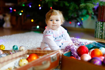 Fototapeta na wymiar Adorable baby girl holding colorful lights garland in cute hands. Little child in festive clothes decorating Christmas tree with family. First celebration of traditional holiday called Weihnachten