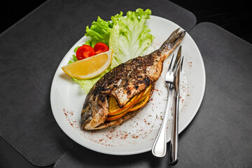 Grilled dorado fish stuffed with orange, garnished with lettuce, lemon and cherry tomato, knife and fork