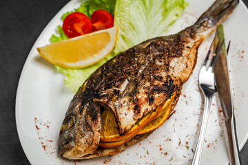 A close-up of grilled dorado fish stuffed with orange, garnished with lettuce, lemon and cherry tomato, knife and fork