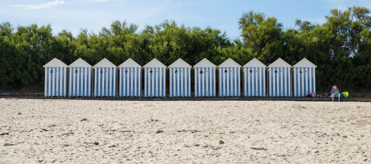 Vieille dame assise à côté d'une rangée de cabanes de plage rayées bleu et blanc au bord de la...