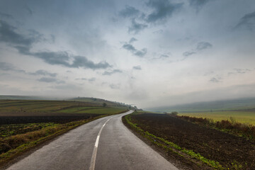 Fototapeta na wymiar Wet asphalt road leading through green agricultural fields at late autumn, fog and thick rain clouds.