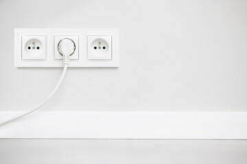 Electrical socket isolated on gray wall. White wire plug plugged in. Renovated studio apartment power supply background. Empty copy space triple white plastic power outlet.