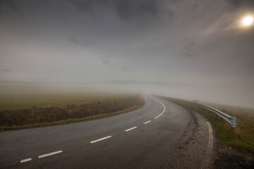 Curving, wet asphalt road in dense fog, sun shining behind thick clouds.