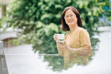 Portrait of smiling mature Asian woman in glasses standing outdoors with cup of coffee