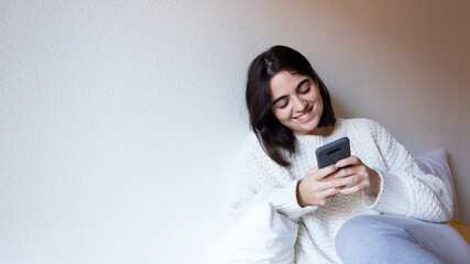 Naklejka premium Woman indoor portrait. Young beautiful using her mobile phone. woman in warm knitted clothes at home. fashion. Autumn, winter
