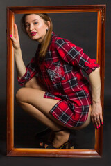 Studio photo of a young woman with long brown hair in a red checked shirt and bare legs. Squatting in a wooden picture frame