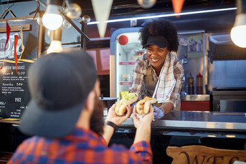 young afro-american employee giving with smile sandwiches through a fast food window to a satisfied customer - 387739573