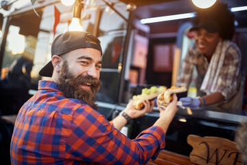 happy, satisfied caucasian beardy male customer taking  sandwiches from a polite employee in fast food service, looking at camera, laughing