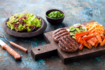 Grilled burger cutlets with baked sweet potato, vegetables and salad served on wood chopping board,...