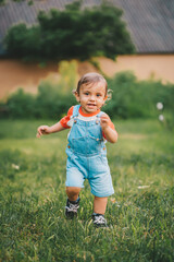 Cheerful perky toddler boy runs to meeting to camera with smile and happiness. Baby boy in denim overall in green park or garden.