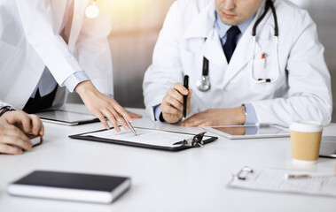 Professional middle aged doctor is talking to his colleagues, while sitting at the desk in a sunny cabinet at hospital. Team of doctors discussing blood sample's results in a clinic. Medicine and