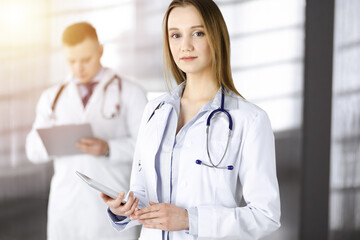 Professional woman doctor with a stethoscope is using a computer tablet, while she is standing together with her colleague in a sunny clinic. Young doctors at work in a hospital. Medicine concept