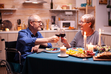 Handicapped man having dinner with man in kitchen toasting. Wheelchair immobilized paralyzed handicapped man dining with wife at home, enjoying the meal