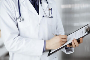 Unknown male doctor standing and working with clipboard of medication history records in clinic at his working place, closeup. Perfect medical service in hospital. Medicine and healthcare concept