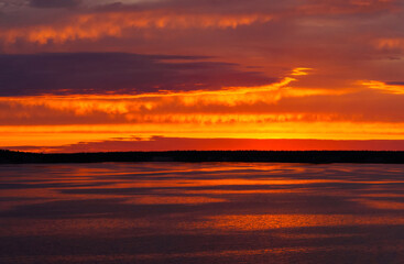 Fototapeta na wymiar A fiery sunset is reflected in the calm river water, overhanging clouds in the bright orange sky, light waves on the water surface.